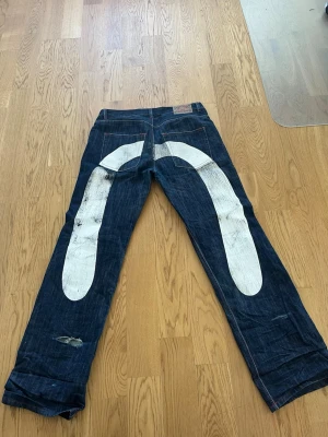 Evisu jeans med vit målad båge - Unika mörkblå jeans från Evisu med den ikoniska vita handmålade bågen på baksidan och en mindre båge på framsidan. Klassisk femficksmodell i kraftig denim med raka ben och röda sömmar. Perfekta för dig som vill sticka ut med streetwear-vibe.