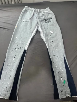 Grå mjukisbyxor från Gallery Dept. - Jag har aldrig använd byxorna‼️‼️. Säljer ett par grå mjukisbyxor från Gallery Dept. med svart och vit panel längs benen och färgstänk som ger en unik streetwear-look. Byxorna har resår i midjan med dragsko och tryckt logga på vänster sida. Perfekta för dig som gillar trendig och avslappnad stil.