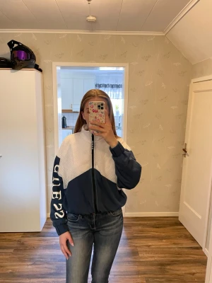 Blåvit vindjacka med tryck på ärmen - Snygg vindjacka i blått och vitt med dragkedja framtill och resår i midja och ärmslut. Jackan har ett coolt tryck på ena ärmen och meshdetaljer upptill. Perfekt för dig som gillar sportig stil och vill sticka ut lite extra.