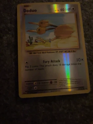 Doduo Pokémonkort holografiskt - Säljer ett holografiskt Doduo Pokémonkort med guldkant. Kortet har motiv av Doduo i en ökenmiljö och text på engelska. Baksidan har klassiskt Pokémon-tryck med Pokéball. Perfekt för samlare eller Pokémonfans.