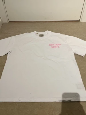Vit Gallery Dept. t-shirt rosa tryck - Vit t-shirt från Gallery Dept. med rosa tryck fram och bak. Klassisk rund hals och normal passform. Materialet är mjuk bomull och trycket fram visar 'GALLERY DEPT.' medan ryggen har större text med 'GALLERY DEPT. HOLLYWOOD, CA.'. Perfekt för dig som gillar streetwear.