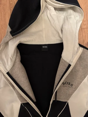 Svart, grå och vit hoodie från BOSS - Snygg hoodie från BOSS med dragkedja, huva och färgblock i svart, grått och vitt. Märkeslogga på bröstet och cool design med kontrasterande paneler. Perfekt för dig som gillar streetwear och vill ha något stilrent men ändå sportigt.