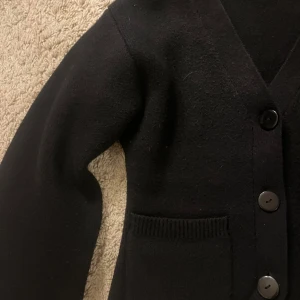 Svart kofta med knappar - Stilren svart kofta med v-ringning och stora knappar framtill. Två rymliga fickor framtill och ribbade detaljer vid fickorna. 