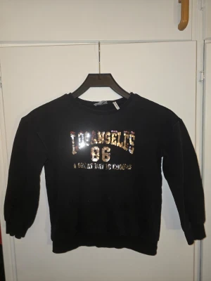 Svart sweatshirt Koton Kids Los Angeles - Cool svart sweatshirt från Koton Kids med glittrande paljetttext 'Los Angeles 86' på bröstet. Tröjan har rund hals, långa ärmar och ribbade muddar. Perfekt för dig som gillar streetstyle och vill sticka ut lite extra.