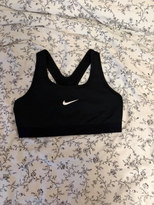 Svart sport-bh från Nike - Nike Swoosh sport-bh med vit logga framtill. Bh:n har brottarrygg och bred resår under bysten för  stöd. Tillverkad i stretchigt funktionsmaterial som passar perfekt till träning och aktivitet. Strl 146-156 men passar mig som har xs-s. Frågor eller mått, tveka inte💗 Nypris: ~320kr. Kan ev diskutera pris