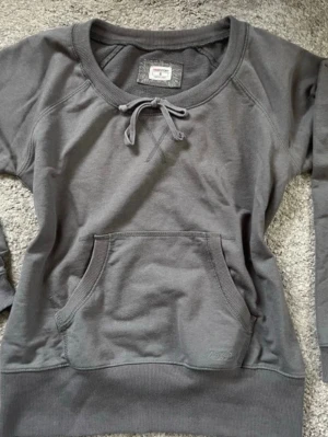 Grå sweatshirt från H&M Sport - Mjuk grå sweatshirt från H&M Sport med rund halsringning och dekorativ knytning framtill. Tröjan har en stor magficka och ribbade muddar vid ärmslut och nederkant. Tillverkad i bomullsmix för en bekväm känsla.