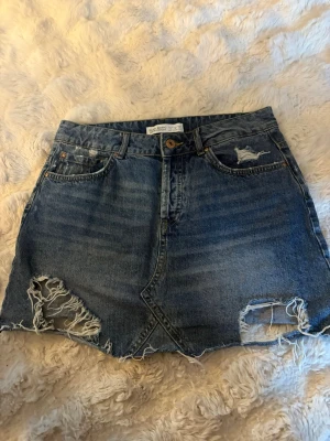Blå jeanskjol med slitningar - Säljer en blå jeanskjol från Zara i storlek M. Kjolen är mellanlång och kan användas både låg och hög midjad. Super snygga slitna detaljer!💕💕💕