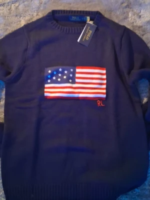 Ralph lauren USA flag - Mörkblå stickad tröja från Polo Ralph Lauren med amerikansk flagga broderad på bröstet och RL-detalj i rött. Klassisk rund halsringning och ribbade muddar. Perfekt för dig som vill ha en ikonisk och stilren look.