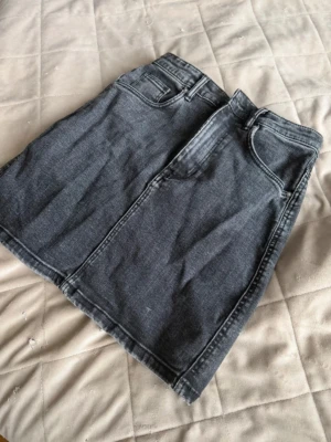 Svart jeanskjol med fickor - Snygg svart jeanskjol i klassisk modell med fem fickor och hällor för bälte. Kjolen har en rak passform och är tillverkad i denim. Perfekt att matcha med en t-shirt eller hoodie för en avslappnad look.
