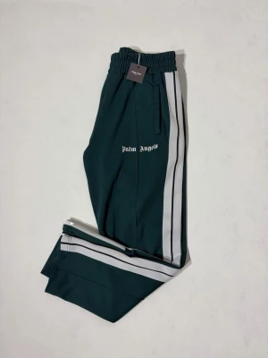 Palm Angels Track Pants - Mörkgröna byxor från Palm Angels i fint skick överlag. Tyvärr har storlekslappen rivits av men Certilogo finns kvar för äkthetsverifiering. En liten reva på höger ben vilket endast syns vid noga inspektion. 