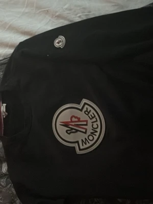 Svart sweatshirt från Moncler - Säljer en svart sweatshirt från Moncler med stort, ikoniskt Moncler-märke på bröstet och en mindre logga på ärmen. Tröjan har rund hals och är tillverkad i mjukt material, perfekt för en stilren och avslappnad look.