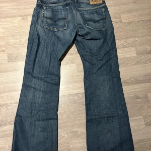 Blå Diesel Larkee jeans bootcut - Säljer ett par blå Diesel Larkee jeans med bootcut passform. Jeansen har klassiska fem fickor, snygga slitningar framtill och en mörk tvätt. Tillverkade i slitstarkt bomullsmaterial och har detaljerade sömmar samt läderpatch bak i midjan.