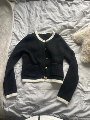 Svart kofta med vita detaljer och guldknappar - Stilren svart kofta från Zara med vita kontrastkanter och snygga guldknappar framtill. Kortare modell med rund halsringning och två fickor fram. Perfekt för att lyfta vilken outfit som helst.