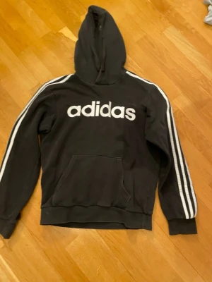 Svart Adidas hoodie med vita ränder - Säljer en svart hoodie från Adidas med klassiska vita ränder längs ärmarna och stor vit logga på bröstet. Hoodien har huva och en stor magficka framtill. Perfekt för en sportig och avslappnad stil.