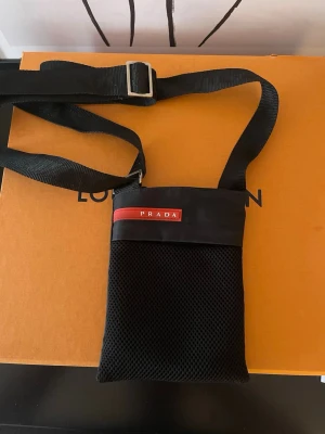 Svart mesh axelväska  - Snygg svart axelväska  med justerbar rem och meshmaterial på framsidan. Väskan har en röd logotypdetalj och dragkedja upptill. Perfekt för att bära det viktigaste och ger en clean streetwear-look.