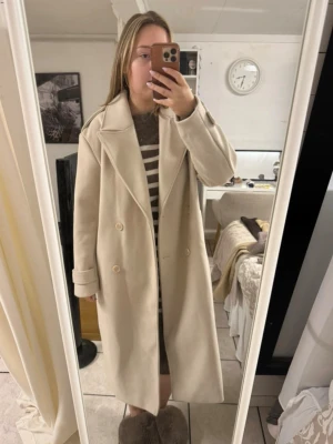 Beige lång kappa med bred krage - Säljer en stilren beige kappa med dubbelknäppning och bred krage. Kappan har långa ärmar med dekorativa passar även M 