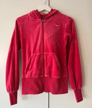 Röd velourhoodie från Nike - Snygg röd hoodie i mjuk velour från Nike med dragkedja framtill, huva och känguruficka. Liten vit Nike-logga på bröstet och breda muddar vid ärmslut och nederkant. Perfekt för en avslappnad och sportig stil.