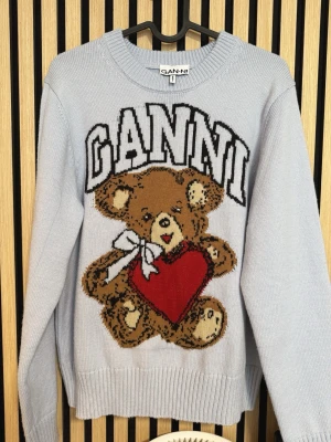 Ljusblå stickad Ganni-tröja med nalle - Superfin ljusblå stickad tröja från Ganni med ett stort nallemotiv som håller ett rött hjärta och en vit rosett. Tröjan har rund halsringning och ribbade muddar. Perfekt statement-plagg för dig som gillar lekfull stil. 