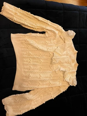 Krämvit stickad tröja med volang - Säljer en krämvit stickad tröja med dekorativa volanger och spetsdetaljer vid halsen och ärmsluten. Tröjan har ett unikt mönster med små bollar och ribbstickade muddar. Perfekt för dig som gillar romantisk och trendig stil.
