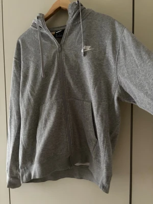 Grå zip hoodie från Nike - Säljer en klassisk grå zip hoodie från Nike med vit logga på bröstet. Hoodien har dragkedja, två stora fickor fram och justerbar huva med snören. Tillverkad i mjuk bomullsblandning som är riktigt skön att ha på sig. Perfekt till chill eller streetwear. Nypris 700kr