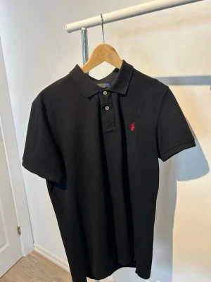 Svart pikétröja från Polo Ralph Lauren - Klassisk svart pikétröja från Polo Ralph Lauren med korta ärmar och krage. Tröjan har knappar framtill och den ikoniska röda loggan broderad på bröstet. Tillverkad i bomull för en skön och stilren look.