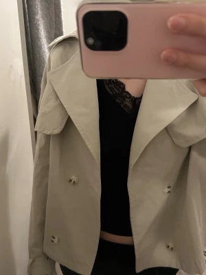 Kort beige trenchcoat  - Snygg kort trenchcoat i beige med dubbelknäppning och stora slag. Jackan har axelklaffar och bälte vid ärmsluten för extra detalj. Tillverkad i ett lätt tyg, perfekt för lager på lager och trendig look.