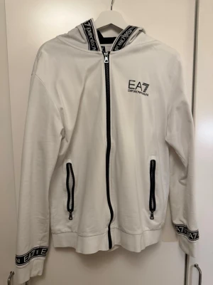 Vit hoodie från Emporio Armani EA7 - Snygg vit hoodie från Emporio Armani EA7 med dragkedja framtill och svarta detaljer. Logga på bröstet och tryck på huva och ärmslut. Två fickor med dragkedja och ribbade muddar. Perfekt för en clean och sportig look. Väldigt liten i pass formen hade nog sagt att den passar M även