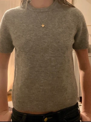 Grå stickad t-shirt i ullblandning - En enkel och stilren grå stickad t-shirt med korta ärmar och rund halsringning. Tröjan är gjord i mjuk ullblandning och passar perfekt till jeans eller kjol för en avslappnad look.