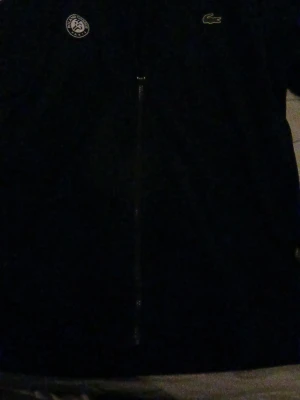 Mörk blå ziptröja Lacoste Roland Garros - Snygg mörk blå ziptröja från Lacoste x Roland Garros-kollektionen. Tröjan har dragkedja framtill, diskret Roland Garros-märke på bröstet och klassiska Lacoste-loggan. Perfekt för tennis eller chill. Materialet känns mjukt och sportigt.