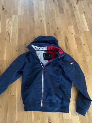 Marinblå vindjacka Tommy Hilfiger - Snygg marinblå vindjacka från Tommy Hilfiger med huva och dragkedja. Jackan har vita och röda detaljer, logga på bröstet och innerkrage med tydlig Tommy Hilfiger-text. Tillverkad i lätt och tåligt syntetmaterial, perfekt för blåsiga dagar.      Storlek 176 sitter som S.  Skriv gärna vid funderingar.   Pris kan diskuteras vid snabb affär 