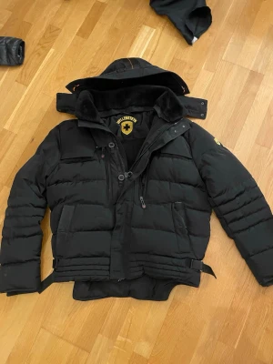 Svart pufferjacka från Wellensteyn - Säljer en svart pufferjacka från Wellensteyn med avtagbar huva och flera praktiska fickor. Jackan har quiltad design, justerbara band i midjan och en tydlig logotyp på insidan och ärmen. Perfekt för kalla vinterdagar och riktigt varm.