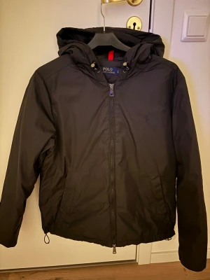Svart vindjacka från Polo Ralph Lauren - Snygg svart vindjacka från Polo Ralph Lauren med huva och dragkedja framtill. Jackan har två sidofickor, justerbar nederkant med dragsko och diskret logga på bröstet. Tillverkad i ett lätt och vindtätt material, perfekt för blåsiga dagar.