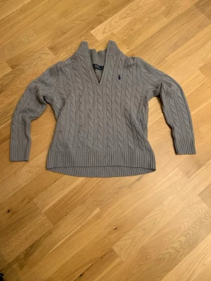 Grå kabelstickad tröja Polo Ralph Lauren - Kabelstickad grå tröja från Polo Ralph Lauren med v-ringning och ribbade muddar. Tröjan har det klassiska mörkblå logobroderiet på bröstet och är tillverkad i mjukt garn. Perfekt för en stilren och avslappnad look.