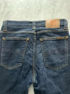 Mörkblå jeans från Nudie Jeans - Snygga mörkblå jeans från Nudie Jeans med klassisk femficksmodell och kontrastsömmar. Jeansen har en rak passform och är tillverkade i slitstark denim av bomull. Nudie-logga på bakfickan och patch i läder baktill i midjan. Modell High kai.