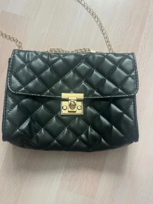 Svart quiltad axelväska med kedja - Snygg svart axelväska i quiltat mönster med guldfärgad kedjerem och guldfärgat vridlås. Väskan har en klassisk rektangulär form och är tillverkad i konstläder. Perfekt accessoar för att lyfta din outfit.