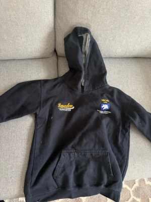 Svart Sweden Supporter hoodie  - Svart hoodie med huva från Sweden Supporter. Tryck i gult och blått på bröstet med texten 'Sweden Supporter' och en logga med tre kronor. Stor magficka framtill. Tillverkad i 80% bomull och 20% polyester. Perfekt för dig som vill visa ditt stöd för Sverige.