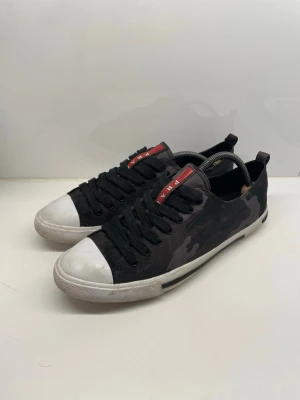 Prada skor  - Snygga svarta sneakers från Prada med kamouflagemönster och vita tåhättor. Skorna har svarta skosnören, låg profil och en röd logga på plösen. Yttermaterialet är textil och gummi, vilket ger en cool och urban look.