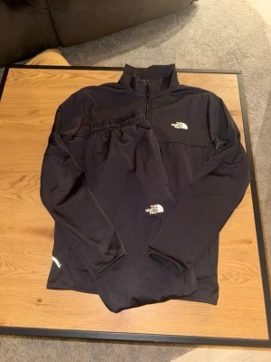 Svart träningsset från The North Face - Säljer ett svart träningsset från The North Face med jacka och byxor. Jackan har dragkedja, hög krage och vita logotyper på bröst och ärm. Byxorna har elastisk midja och logotyp på benet. Perfekt för sport och träning.