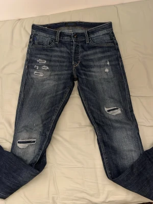 Blå slitna jeans från Jack & Jones - Säljer ett par mörkblå jeans från Jack & Jones med slitningar och hål på både fram- och baksida. Jeansen har klassisk femficksmodell, raka ben och snygga detaljer på bakfickorna. Perfekta för en avslappnad och trendig look. Stl 29/32