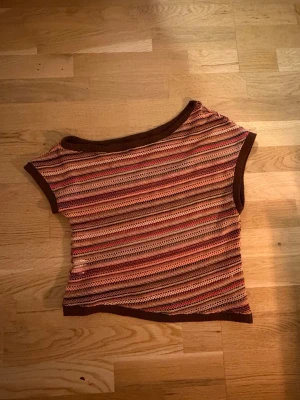 Randig ribbad topp i bruna toner - Säljer en kortärmad, ribbad topp med randigt mönster i bruna, orangea och rosa nyanser. Toppen har en asymmetrisk halsringning och är gjord i ett genombrutet, stickat material som ger en luftig känsla. Perfekt för dig som gillar retrovibbar och färg!