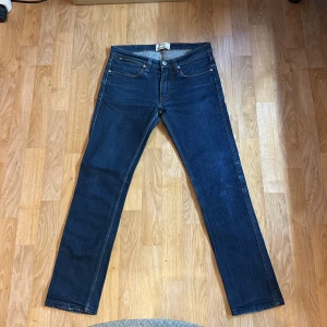 Mörkblå slimfit jeans från Acne Studios - Snygga mörkblå jeans från Acne Studios med rak passform och klassisk femficksdesign. Jeansen har orange kontrastsömmar och metallknapp framtill. Perfekta för en clean och stilren look. Materialet är denim i bomull.