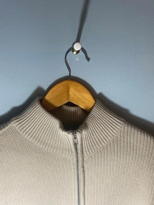 Beige stickad ziptröja med hög krage - Beige stickad tröja med halvzip och hög krage. Ribbad struktur och slim fit-passform. Fint skick utan synliga skador – perfekt som lager på lager eller att bära som den är.