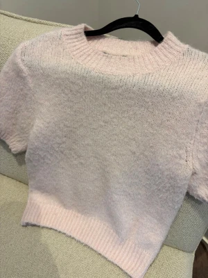 Ljusrosa stickad kortärmad tröja - Supersöt ljusrosa stickad tröja med korta ärmar och bred ribbad kant nedtill samt vid halsen. Tröjan har en mjuk och fluffig känsla, perfekt för lager på lager. Passar dig som gillar en enkel men trendig look.