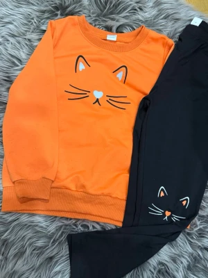 Orange och svart kattset - Säljer ett gulligt set med orange långärmad tröja och svarta byxor, båda med tryck av en söt kattansikte. Tröjan har ribbade muddar och rund hals. Perfekt för dig som gillar djurmotiv och vill ha något bekvämt och lekfullt.