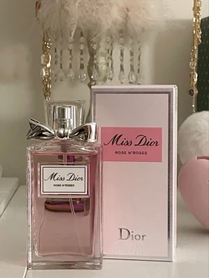 Miss Dior Rose N'Roses parfym - Miss Dior Rose N'Roses är en elegant parfym från Dior med en ljusrosa vätska i en rektangulär glasflaska. Flaskan har en silverfärgad metallkork med en dekorativ rosett. Kartongen är ljusrosa med rosa detaljer och klassisk Dior-logga.