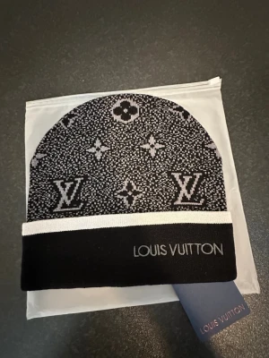Svart mössa från Louiss Vuitton - Säljer en svart och grå mössa från Louiss Vuitton med klassiskt monogrammönster och vit kant. Mössan har broderad logotyp framtill och är tillverkad i mjukt stickat material. Perfekt accessoar för dig som vill ha en exklusiv och trendig look.Följ tt:Marknads.closet