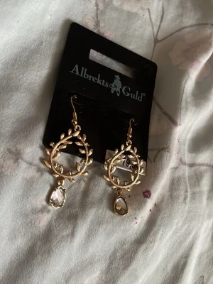 Guldörhängen med kristall från Albrekts Guld - Snygga örhängen i guldfärgad metall från Albrekts Guld. De har en öppen cirkelform med bladliknande detaljer och en klar kristallpärla som hänger nedtill. Perfekta för dig som gillar unika accessoarer med elegant touch.