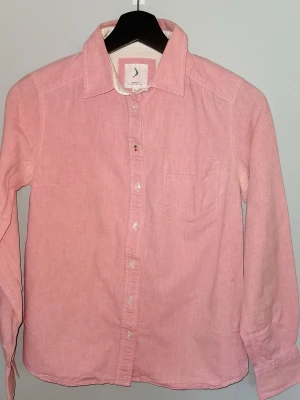 Fin rosa Oxfordskjorta i bomull med ficka från Boomerang - Snygg rosa skjorta i bomull med klassisk krage och bröstficka. Skjortan har långa ärmar och knäppning framtill. Perfekt för en avslappnad men stilren look. Har den typiska Boomerang-loggan i ena knappen. Från märket Boomerang storlek 34. Är i använd skick, något blek i färgen under armarna (se bild) i övrigt fint skick. 