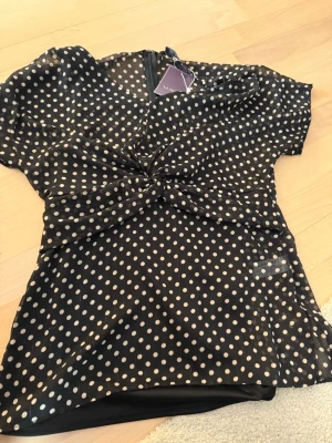 Svart prickig blus från La Mode - Snygg svart blus från La Mode med vita prickar och twistad detalj framtill. Blusen har v-ringning och korta ärmar, tillverkad i ett lätt och luftigt polyestertyg. Perfekt för dig som gillar trendiga mönster och en feminin look.