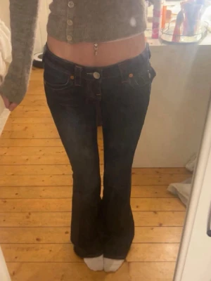 Mörkblå bootcut jeans True Religion - Säljer ett par mörkblå low waist bootcut jeans från True Religion med kontrastsömmar och stora fickor bak med dekorativa knappar. Klassisk femficksmodell och slitstark denim. Perfekta för dig som gillar en retro och trendig look.